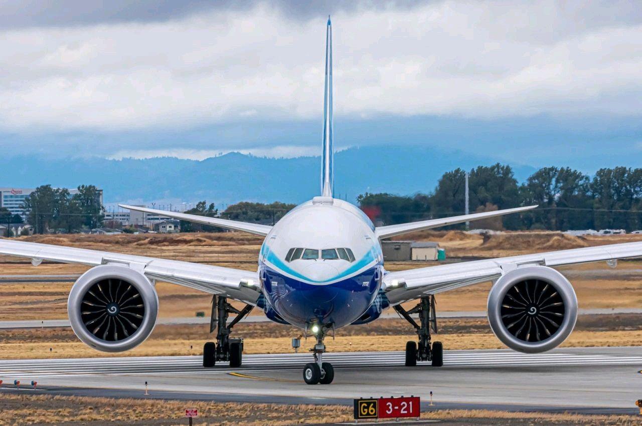 Front view of a commercial airplane on the runway, with mountains and cloudy sky in the background.