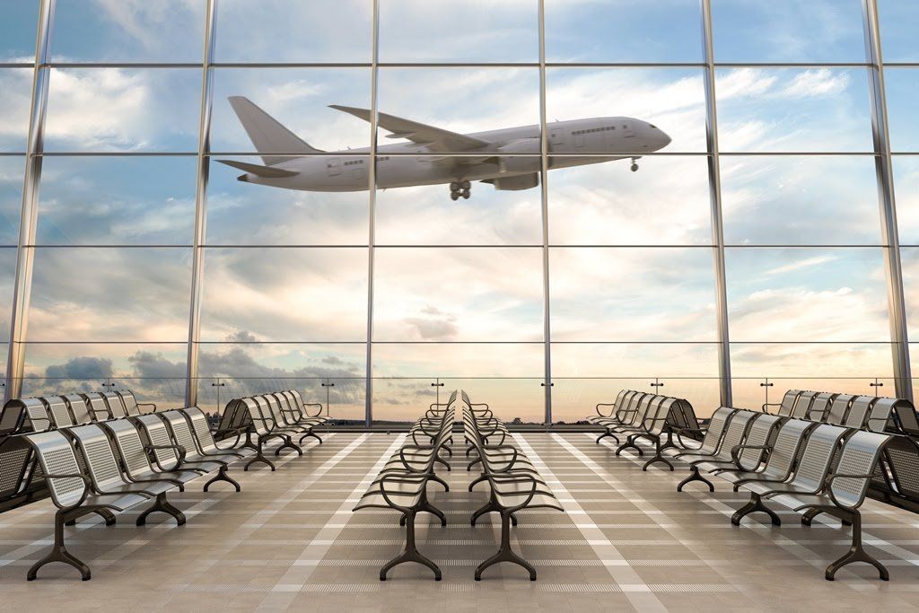 Empty airport terminal seating area with large windows showing a plane taking off against a cloudy sky.